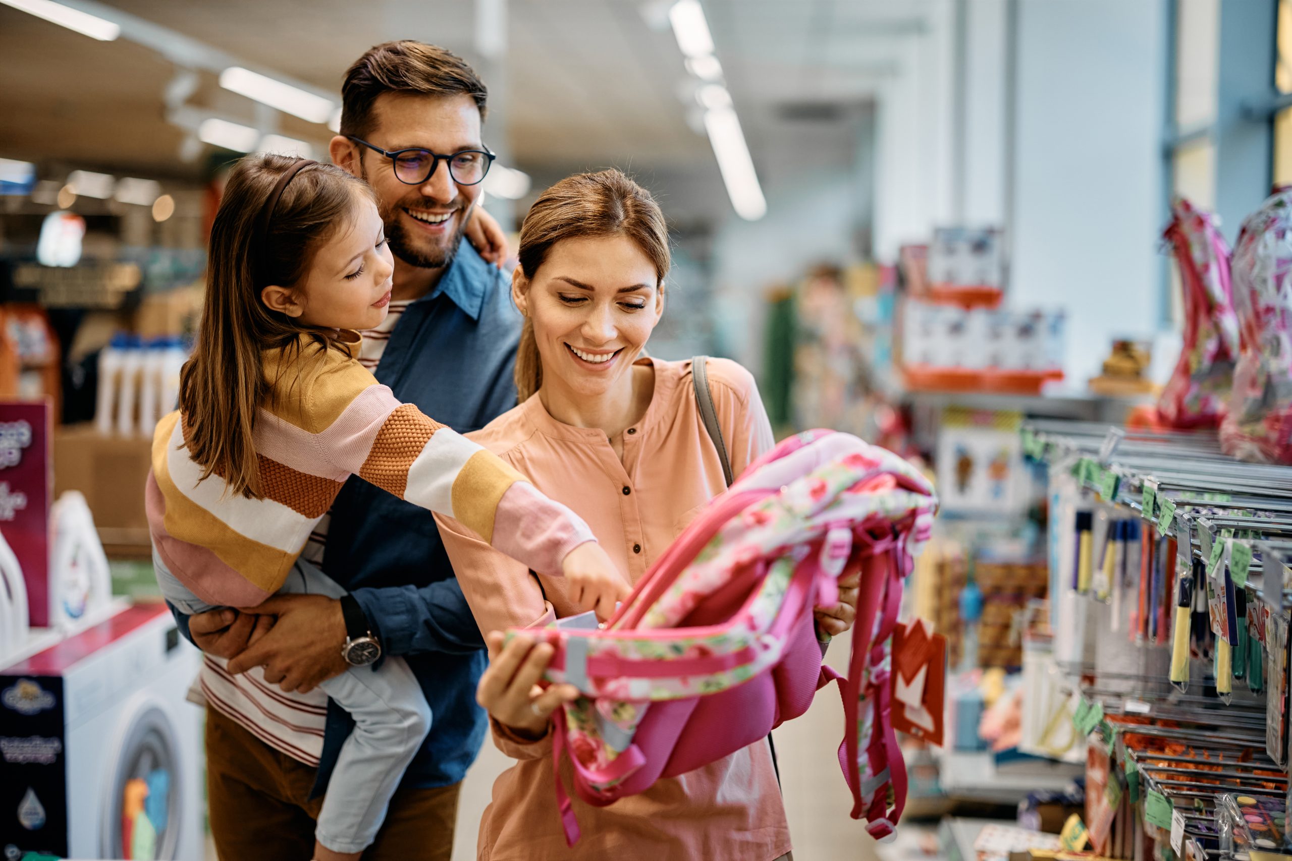 Happy little girl pointing at backpack while buying school supplies with her parents in supermarket.