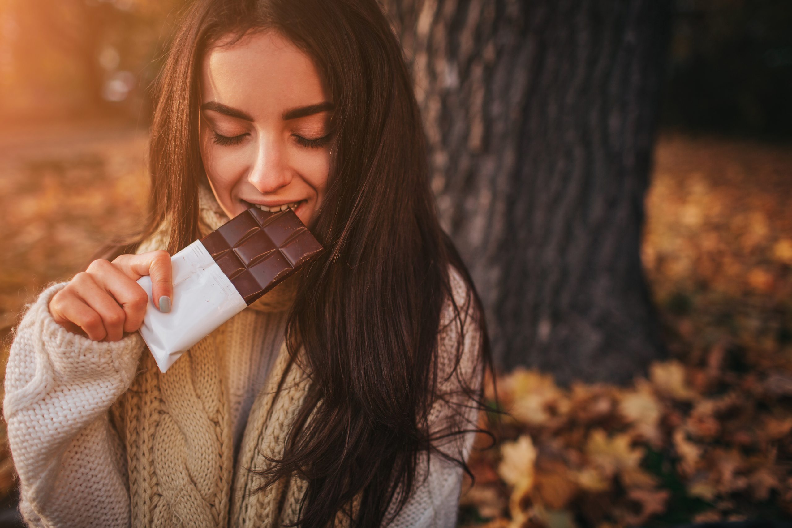 Beautiful young brunette sitting on a fallen autumn leaves in a park, eating chocolate bar